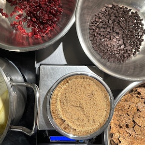 May include: A top-down view of baking ingredients in stainless steel bowls, including dried cranberries, dark chocolate chips, brown sugar on a digital scale, a bowl of broken crackers, and a pot with melting butter sticks on a stovetop.