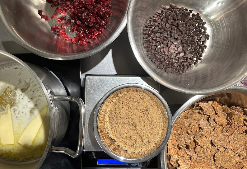 May include: A top-down view of baking ingredients in stainless steel bowls, including dried cranberries, dark chocolate chips, brown sugar on a digital scale, a bowl of broken crackers, and a pot with melting butter sticks on a stovetop.