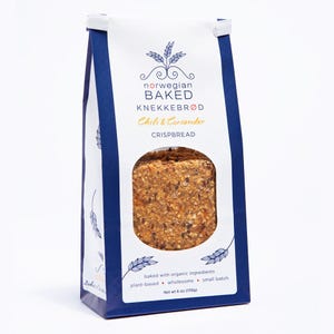 May include: A white and dark blue paper bag of Norwegian Baked Knekkebr&oslash;d Chili & Coriander crispbread. The packaging features a circular window revealing the textured, multi-grain crispbread inside, with text stating net wt 6 oz (170g).