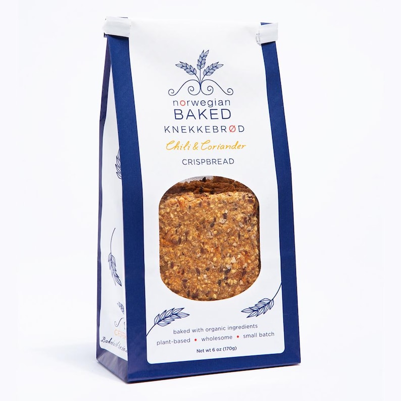 May include: A white and dark blue paper bag of Norwegian Baked Knekkebr&oslash;d Chili & Coriander crispbread. The packaging features a circular window revealing the textured, multi-grain crispbread inside, with text stating net wt 6 oz (170g).