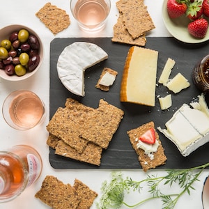 May include: A black slate serving board featuring a selection of cheeses, including a round brie, a wedge of hard cheese, and a soft cheese, accompanied by multi-grain crackers, a jar of fig jam, fresh strawberries, green and black olives, and melon.