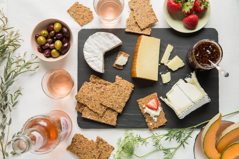 May include: A black slate serving board featuring a selection of cheeses, including a round brie, a wedge of hard cheese, and a soft cheese, accompanied by multi-grain crackers, a jar of fig jam, fresh strawberries, green and black olives, and melon.