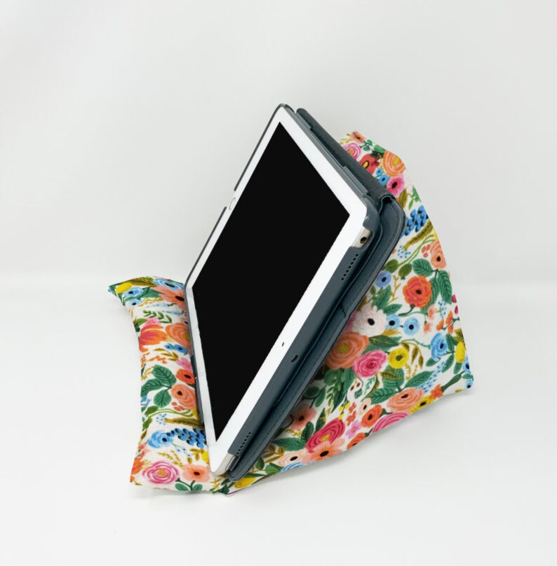 Puede incluir: Un soporte de almohada con estampado floral con una tableta apoyada en &eacute;l. El soporte est&aacute; hecho de tela y tiene una forma triangular. La tableta est&aacute; en una funda negra.