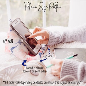 May include: A white and pink floral phone pillow with channel cushions on both sides. The pillow is 5 inches tall and 6.5 inches wide. "Fit may vary depending on device on pillow. This is just an example."