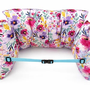 May include: A floral bath pillow with a light blue strap and buckle. The pillow has a white background with pink, purple, and yellow flowers and green leaves. The pillow is designed to support the head and neck.