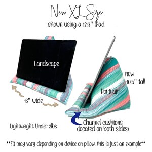 May include: A teal and pink striped pillow with channel cushions on both sides. The pillow is designed to hold a tablet in either landscape or portrait orientation. The pillow is 13 inches wide and 10.5 inches tall. "New XL Size shown using a 12.9" iPad" is written above the pillow. "Landscape" and "Portrait" are written on the pillows. "Lightweight: Under 2lbs" is written below the pillow. "Fit may vary depending on device on pillow. This is just an example" is written below the pillow.