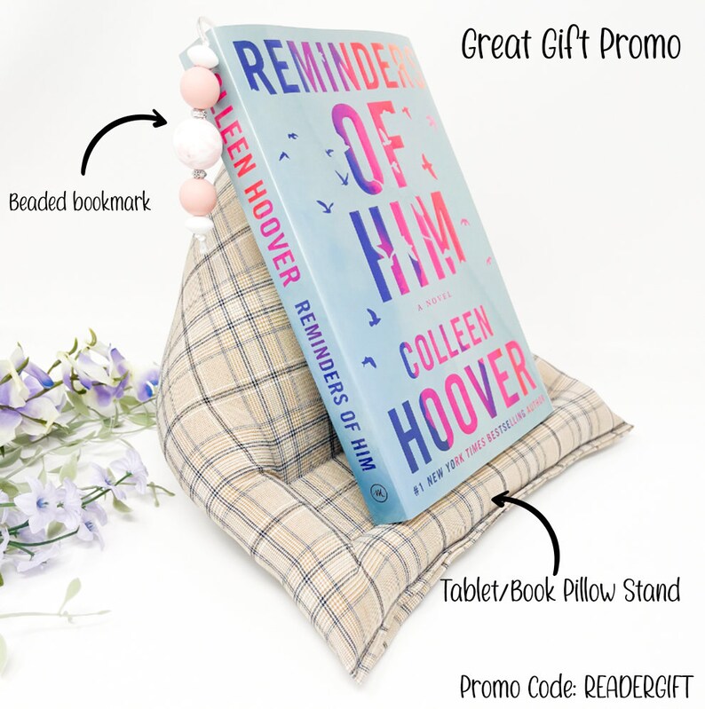 May include: A beige and brown plaid pillow stand with a book titled "Reminders of Him" by Colleen Hoover propped up on it. A beaded bookmark is attached to the book. The text "Great Gift Promo" and "Promo Code: READERGIFT" are also visible.