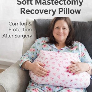May include: A pink and white mastectomy recovery pillow with a bow pattern is held by a person. The pillow is designed for comfort and protection after surgery. The text at the top reads "Soft Mastectomy Recovery Pillow".