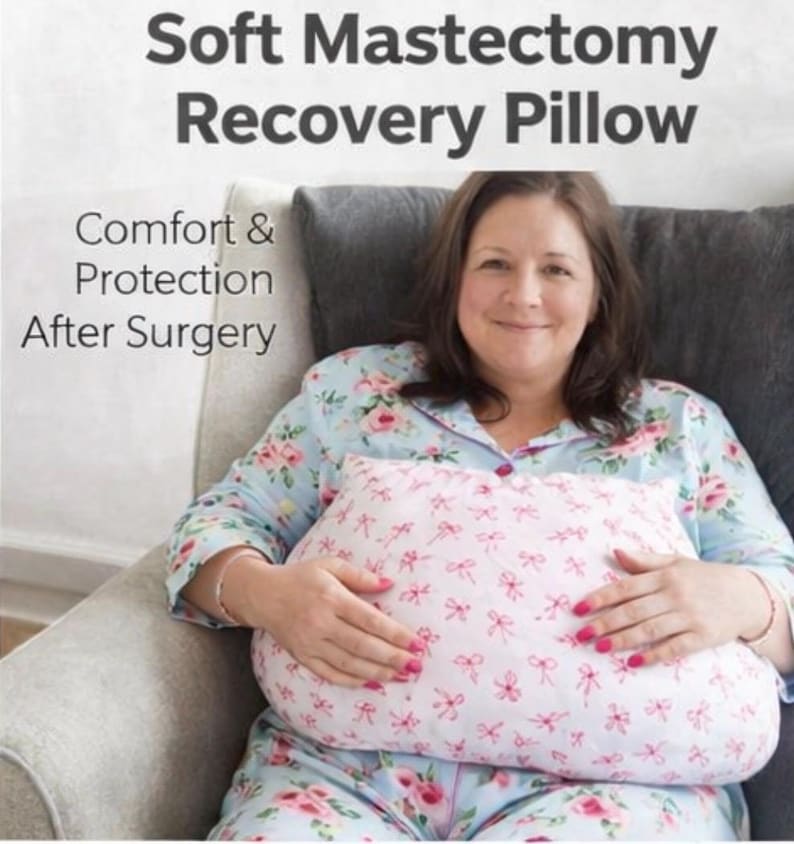May include: A pink and white mastectomy recovery pillow with a bow pattern is held by a person. The pillow is designed for comfort and protection after surgery. The text at the top reads "Soft Mastectomy Recovery Pillow".