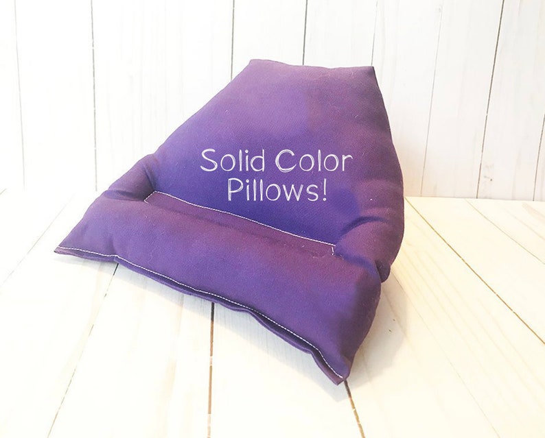 May include: A purple wedge-shaped pillow with a sewn-in pocket for holding a tablet or book. The pillow has white stitching and the text "Solid Color Pillows!" embroidered on the front.