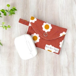 May include: A red fabric pouch with a white daisy pattern and a yellow center. The pouch has a flap closure and a small loop handle. The pouch is sitting on a light wood surface next to a white wireless earbud case. The pouch has a label that says "Etsy Elephant Designs".