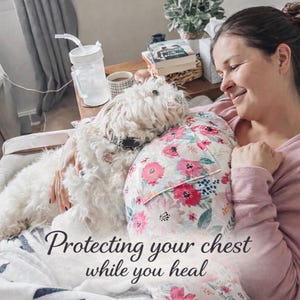 May include: A woman is resting with a white dog and a floral pillow. The pillow has a white background with pink and red flowers. The text "Protecting your chest while you heal" is at the bottom of the image.
