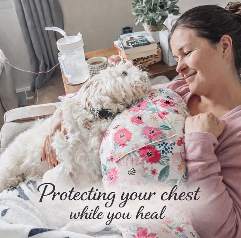 May include: A woman is resting with a white dog and a floral pillow. The pillow has a white background with pink and red flowers. The text "Protecting your chest while you heal" is at the bottom of the image.