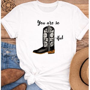Puede incluir: Camiseta blanca con un diseño de bota de vaquero negra adornada con flores blancas. El texto "You are so -iful" está impreso encima de la bota. La camiseta se combina con vaqueros azules, botas marrones y un sombrero de paja.