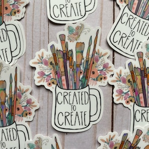 May include: A white coffee mug with the text "Created to Create" in black lettering. The mug is filled with paintbrushes and flowers in shades of pink, orange, and purple.