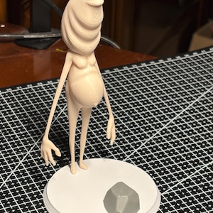 Può includere: Statuetta beige stampata in 3D raffigurante una creatura aliena con una grande testa, piccoli occhi e un ventre incinto. La figura è su una base circolare bianca con una roccia geometrica grigia.