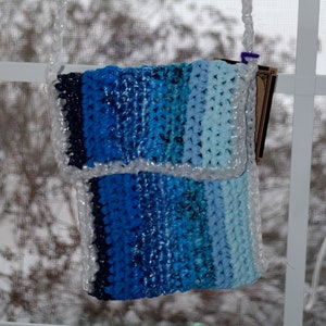 May include: A blue and white crocheted bag with a white plastic strap. The bag is hanging from a window.