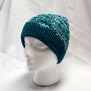 Puede incluir: Un gorro de punto de color azul verdoso y blanco con un patrón texturizado. El gorro está en una cabeza de maniquí blanca.