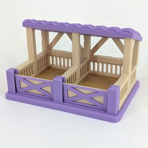 May include: A miniature toy stable with two stalls. The structure features a purple roof and base, with tan-colored walls and supports. The stall doors have an X-shaped design. The interior of the stalls has a light brown floor.