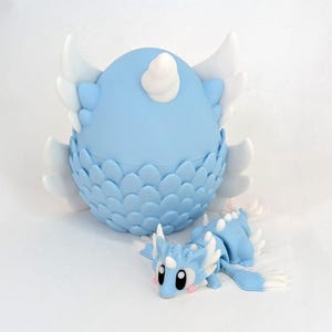 May include: A light blue dragon-themed figurine set. The set includes a large, egg-shaped container with white wings and a small dragon figurine. The container has a scale-like pattern. The dragon figurine is light blue with white accents.