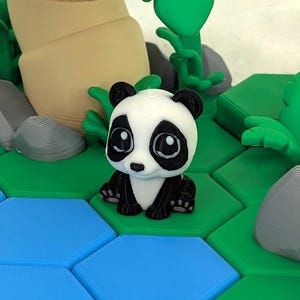 Op de afbeelding: Een 3D-geprinte pandafiguur in zwart en wit, zittend op een groene zeshoekige tegel. De panda wordt omringd door 3D-geprint bamboe en rotsen. De achtergrond heeft blauwe zeshoekige tegels.
