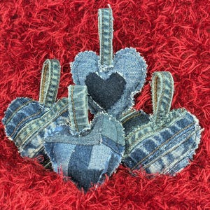 Puede incluir: Cuatro adornos en forma de corazón de mezclilla con lazos para colgar. Los corazones están hechos de varios tonos de mezclilla azul, algunos con parches y una aplicación de corazón azul oscuro. Descansan sobre una superficie roja vibrante y texturizada.