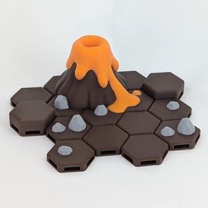 May include: A miniature volcano model with orange lava flowing from the top. The volcano sits on a brown hexagonal tile base with gray rocks scattered around. This model is likely for a board game or diorama.