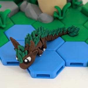 Puede incluir: Una figura de dragón marrón y verde con un diseño detallado. El dragón está posado sobre una baldosa hexagonal azul, con otras baldosas y un fondo de paisaje. La figura tiene una cabeza detallada, cuernos y una larga cola.