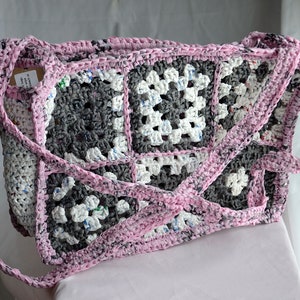 May include: A pink and gray crocheted bag made from recycled plastic. The bag has a square pattern and a long strap.