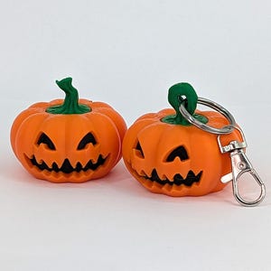 May include: Two orange pumpkin-shaped keychains with carved faces and green stems. One keychain has a silver clasp and ring. The pumpkins have triangular eyes and jagged mouths, perfect for Halloween.