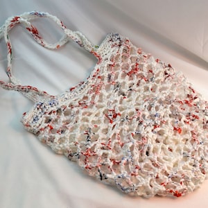 May include: A white crocheted tote bag with red and blue flecks. The bag has a net-like design and two white handles. The bag is made of a durable material and is suitable for carrying groceries or other items.