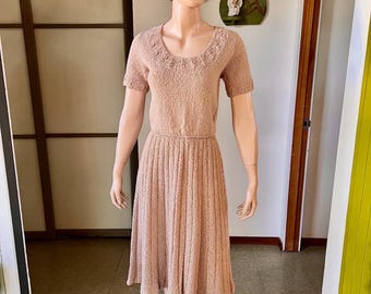 May include: A vintage peach-colored dress displayed on a mannequin. The dress features short sleeves, a scoop neckline with delicate detailing, and a fitted bodice that flows into a pleated skirt. The dress is knee-length and made of a textured knit fabric.