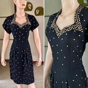 May include: Black vintage dress with gold dot pattern and gold embellishments around the neckline. The dress has short sleeves and a knee-length hemline. The dress is displayed on a mannequin.