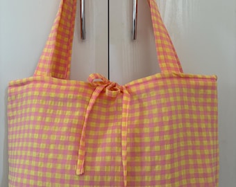 May include: A rectangular fabric tote bag featuring a bright yellow and pink gingham check pattern. The bag includes two shoulder straps and a central fabric tie closure at the top opening, constructed from a lightweight, woven textile material.