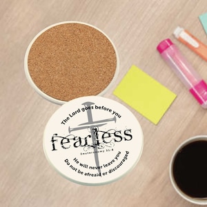 May include: White ceramic coaster with a cork bottom. The coaster features a black and white design of a cross with the words "The Lord goes before you fearless Deuteronomy 31:8 He will never leave you Do not be afraid or discouraged".