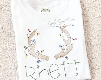May include: White long-sleeved shirt with embroidered reindeer antlers decorated with colorful Christmas lights. The name "Rhett" is embroidered in green below the antlers. The shirt also has the text "Simple Sweet Mama Custom Embroidery".