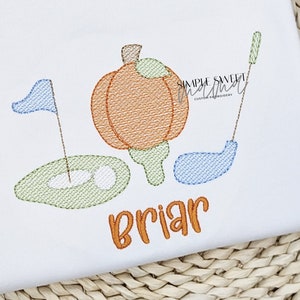 May include: Embroidered design of a pumpkin, golf club, and golf ball on a white fabric with the name "Briar" stitched in orange thread.