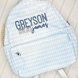 May include: A light blue gingham backpack with a white zipper and a white and blue embroidered name "Greyson" and the text "Saddle Up Sweet Mama James".