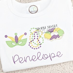 May include: A white t-shirt with purple embroidery of a Mardi Gras mask, beads, and a king cake. The name Penelope is embroidered in purple below the Mardi Gras themed design.