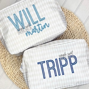 May include: Two personalized cosmetic bags with a light gray and white gingham pattern. One bag has the name "WILL" and "martin" embroidered in blue, and the other has "TRIPP" embroidered in blue. The text "simple sweet MAMA" is also embroidered on both bags.