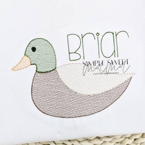 May include: Embroidered design of a brown and green duck with the name "Briar" stitched above it. The design is on a white fabric background. The text "Simple Sweet Stitchal" is also embroidered below the duck.