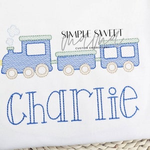 May include: A blue and green embroidered train with a yellow detail and the name "Charlie" stitched below. The train is on a white background.