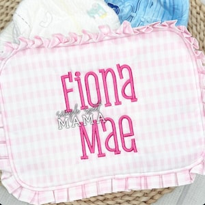 May include: A pink and white gingham baby blanket with the name "Fiona Mae" embroidered in bright pink. The blanket has a ruffled edge and the text "simple sweet MAMA" is also visible.