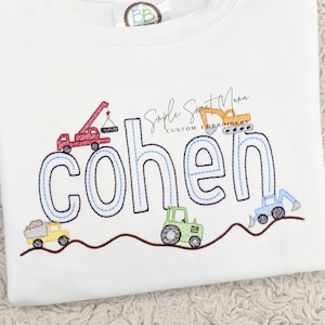 May include: A white t-shirt with the name "Cohen" embroidered in blue outlined letters. The name is surrounded by a brown line and several embroidered construction vehicles, including a crane, a bulldozer, a dump truck, and a tractor.