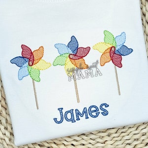 May include: A white t-shirt with three colorful pinwheels embroidered on it. The pinwheels are red, orange, yellow, green, blue, and purple. The name "James" is embroidered in blue below the pinwheels.