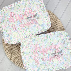 May include: Two floral print diaper and wipe cases with ruffled edges. The cases feature a pastel floral pattern with pink embroidered text that reads "Diapers & Wipes" and "Simply Sweet Mama".