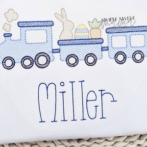 May include: A blue embroidered train with a bunny, Easter eggs, and carrots in the car. The train has three cars and is on a white background. The name "Miller" is embroidered below the train.