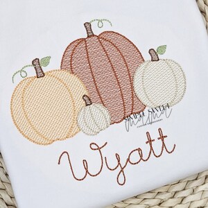 May include: White fabric embroidered with four pumpkins in shades of brown, tan, and white, with green vines. The name "Wyatt" is embroidered in brown script below the pumpkins. The text "SIMPLE SWEET MAMA CUSTOM EMBROIDERY" is also visible.