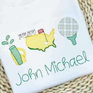 May include: Embroidered design featuring a golf bag, a golf ball, and a green outline of the United States with a red flag. The name "John Michael" is embroidered in green below the design.