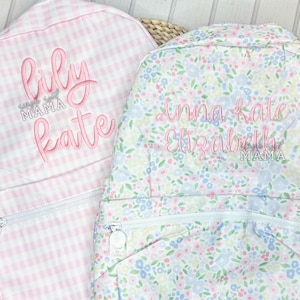 May include: Two pink and white patterned backpacks with embroidered names. The first backpack has the name "lily kate" embroidered on it with the text "mama" below. The second backpack has the name "anna-kate elizabeth" embroidered on it with the text "mama" below.
